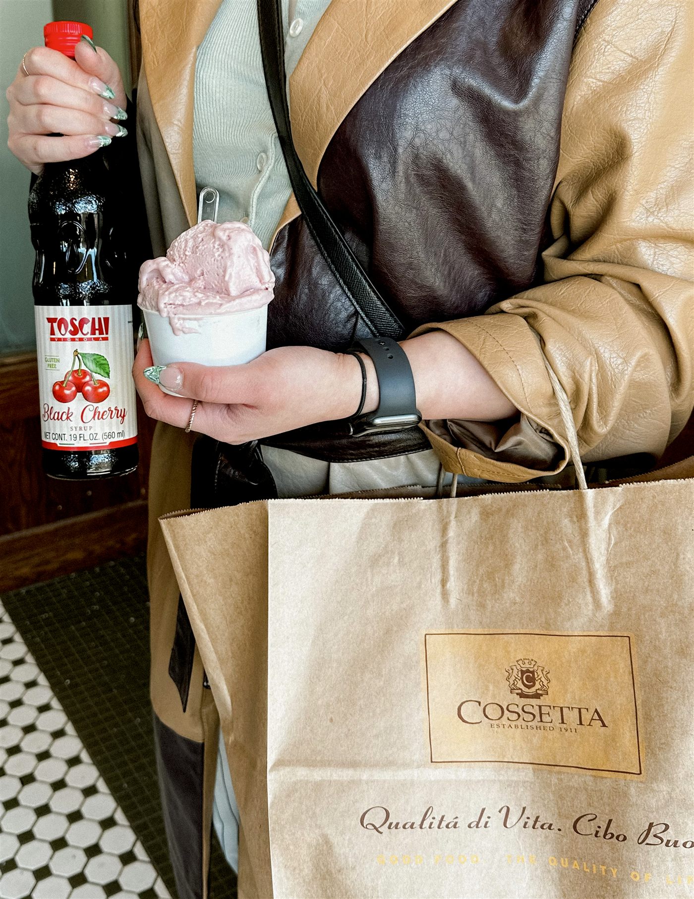A cup of black cherry gelato, a bottle of cherry syrup, and a Cossetta paper bag held in a tan leather coat