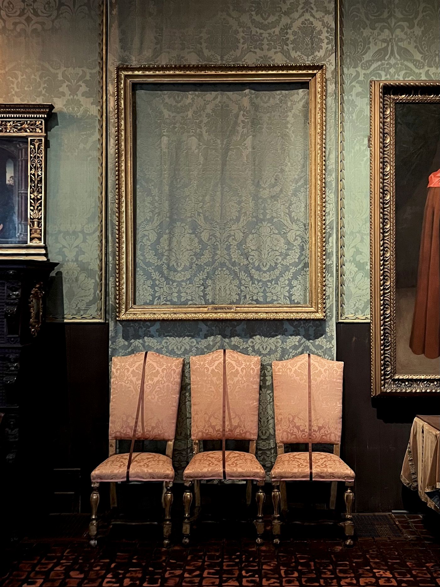 An empty gilt frame on blue-green damask above three pink upholstered chairs