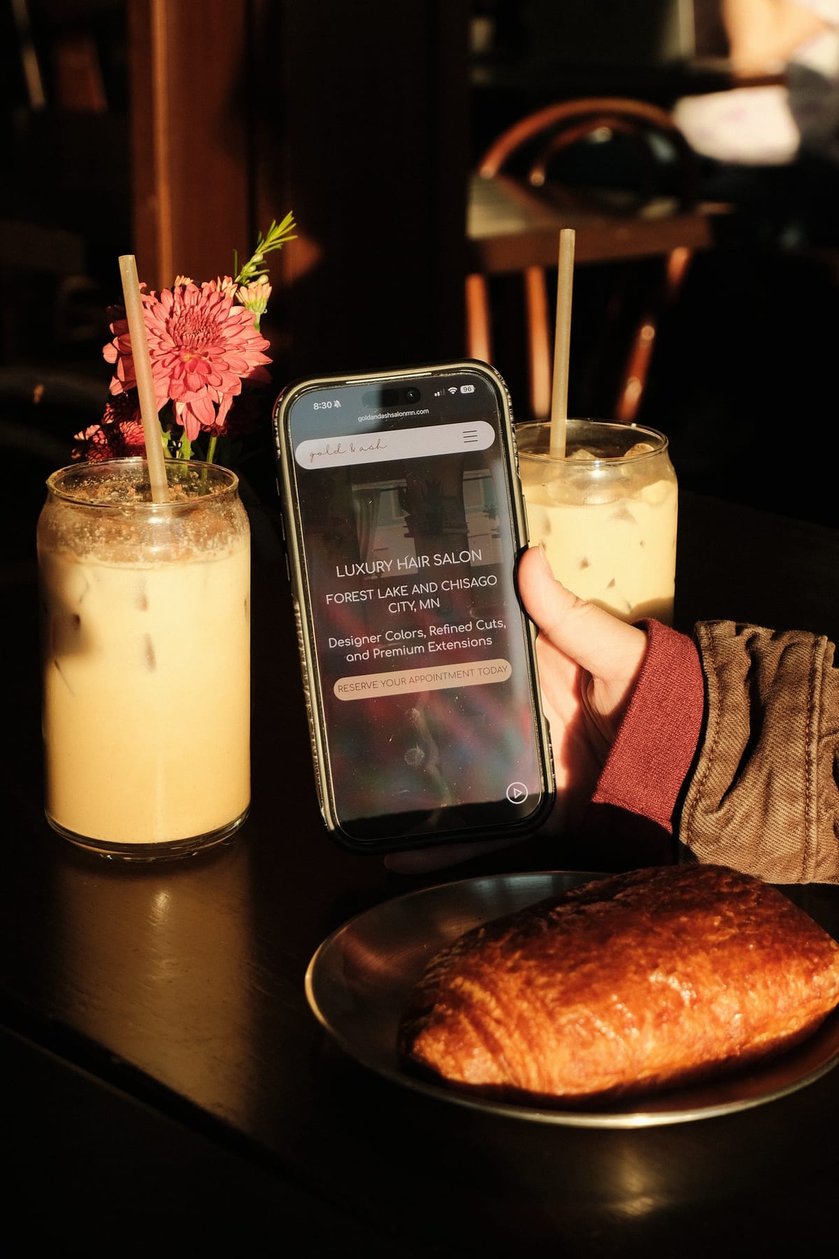 The Gold & Ash website open on a phone beside two iced lattes and a croissant
