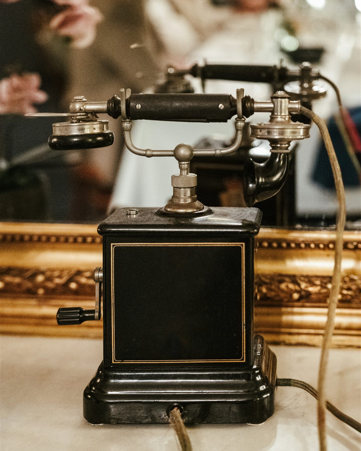 An antique black telephone on a marble shelf before a gilt-framed mirror